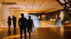 An exhibition space with four figures standing in the space, looking at a lit up moon sculpture.